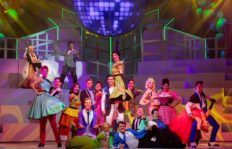 Large ensemble cast in colourful costumes poses on stage beneath a glittering disco ball under purple and golden stage lighting.
