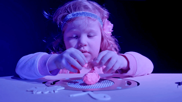 Child wearing a glittery headband concentrates on manipulating pink slime on a surface under blue stage lighting.