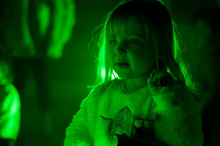 Person with long hair lit by intense green stage lighting, gesturing expressively.