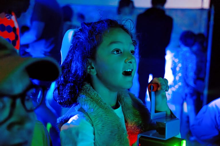 Child's face illuminated by blue and green neon lighting in an interactive installation or exhibition space.