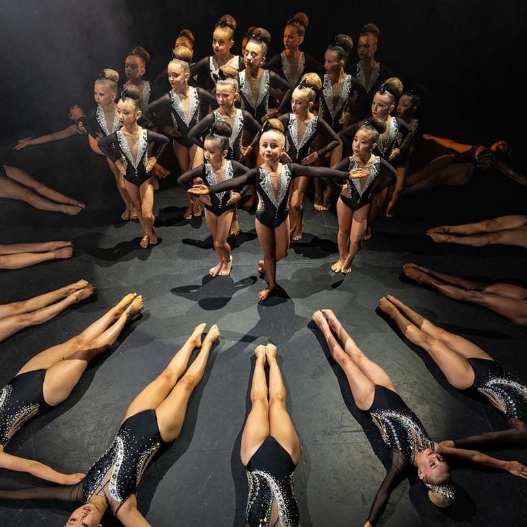 Large ensemble of dancers in formal attire arranged in a circle with legs extended toward centre on dark stage.