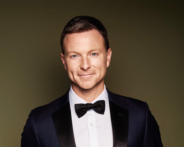 Man in formal black tuxedo and bow tie against neutral green background.