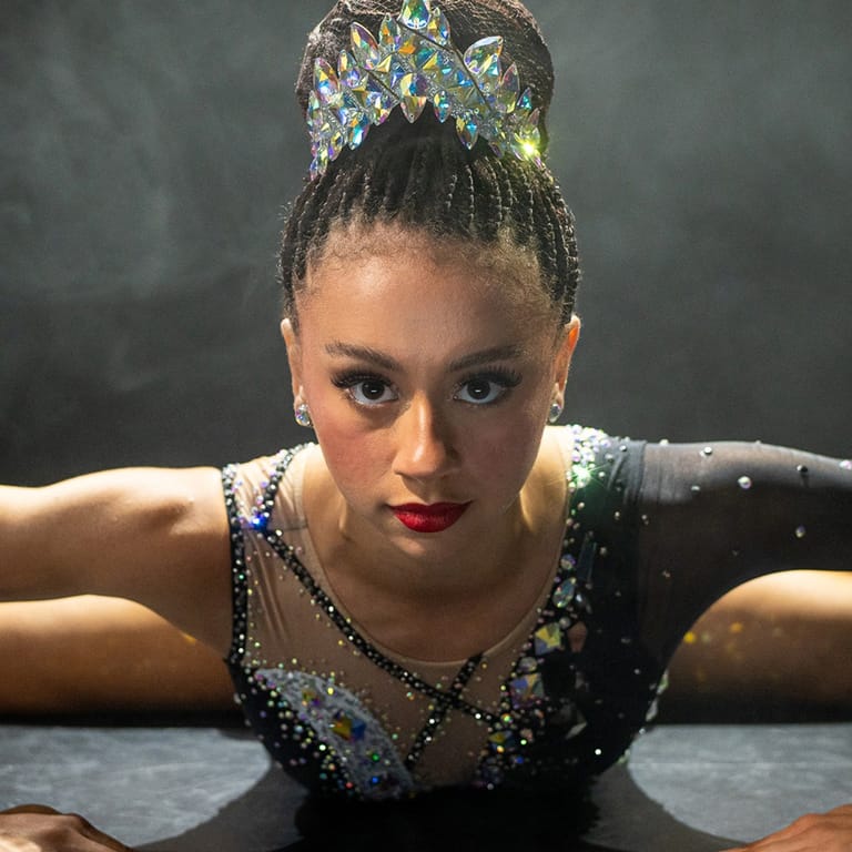 Performer in sequined leotard and jewelled crown poses with arms extended against dark background.