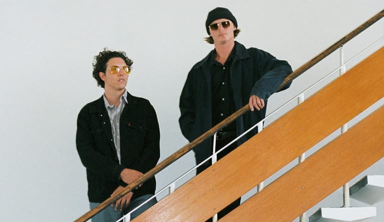 Two people in dark jackets stand on a wooden staircase against a white and orange wall.