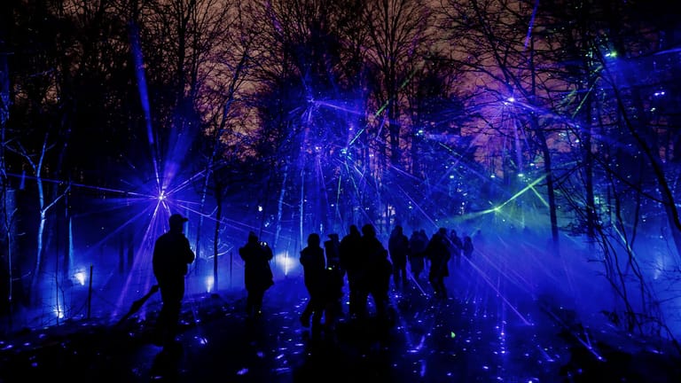 Silhouetted figures walk through an outdoor installation illuminated by blue and purple lights among bare trees.