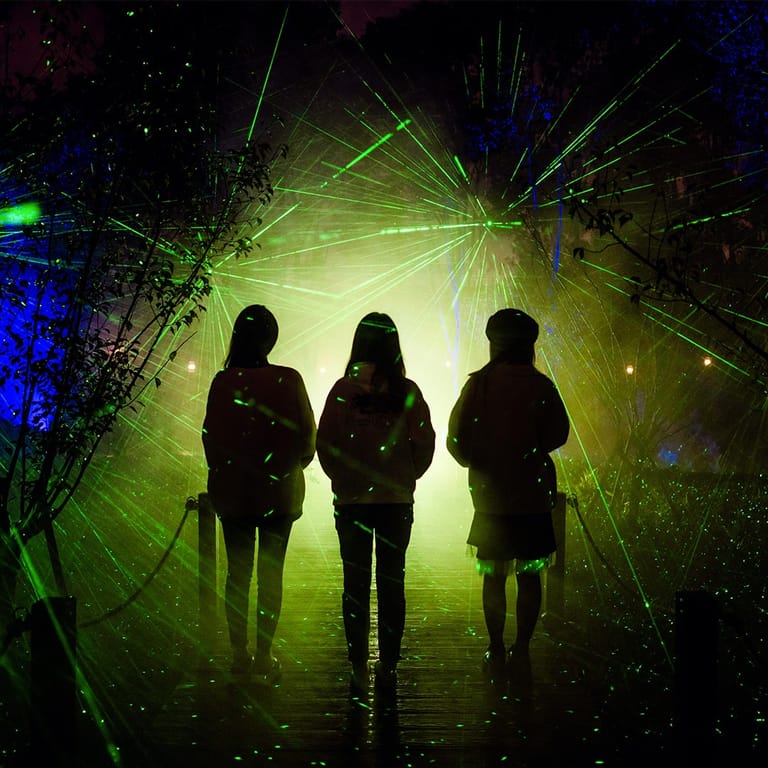 Three silhouetted figures stand amid vivid green and blue laser light effects in a darkened space.