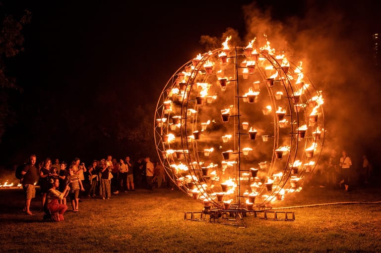 Massive illuminated spherical sculpture glows warmly as silhouetted spectators gather around it at night.