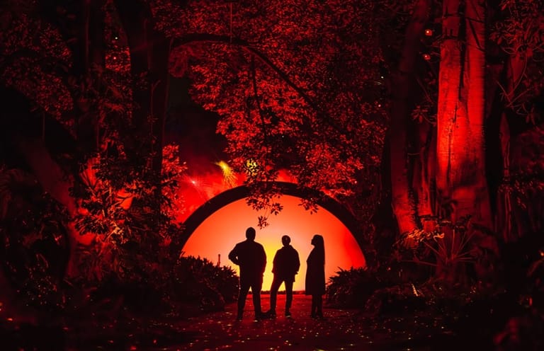 Three silhouetted figures stand beneath a glowing red sun in a darkly textured forest setting.