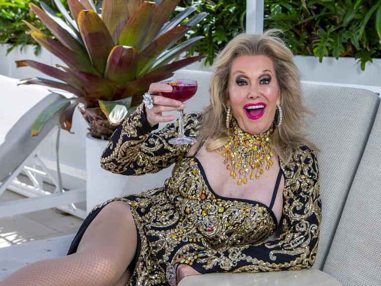 Woman in gold-embellished black outfit holding a cocktail glass, seated outdoors with tropical plants behind.