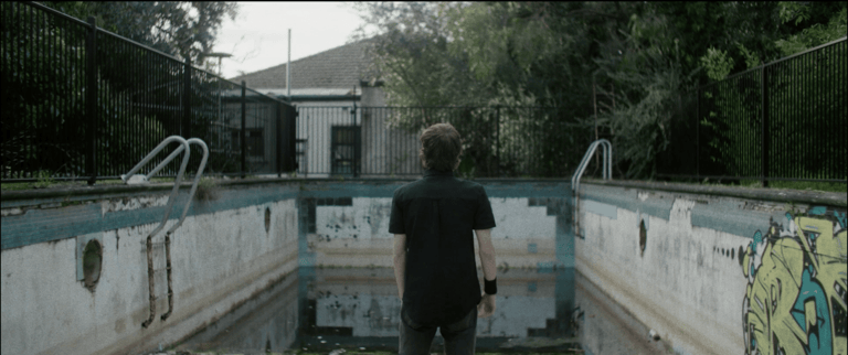 Person in black shirt standing at edge of empty, overgrown swimming pool with weathered concrete walls.