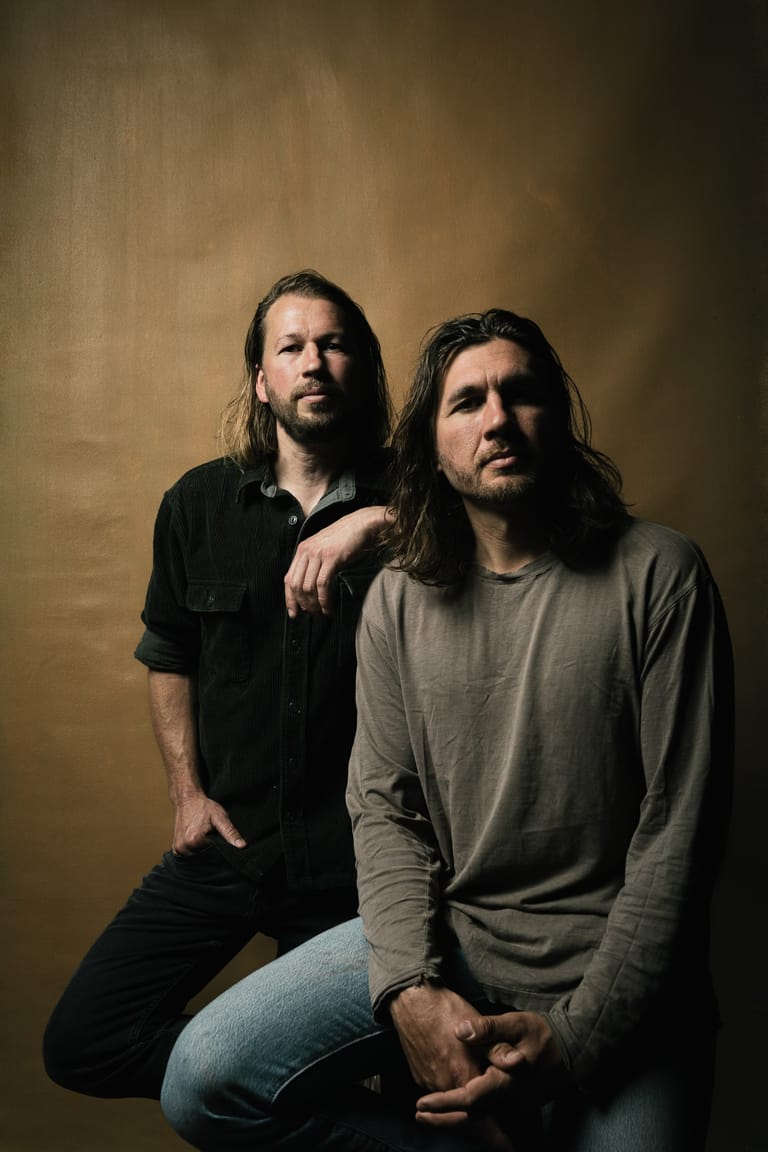 Two men with long dark hair pose against a warm brown backdrop in a studio portrait.