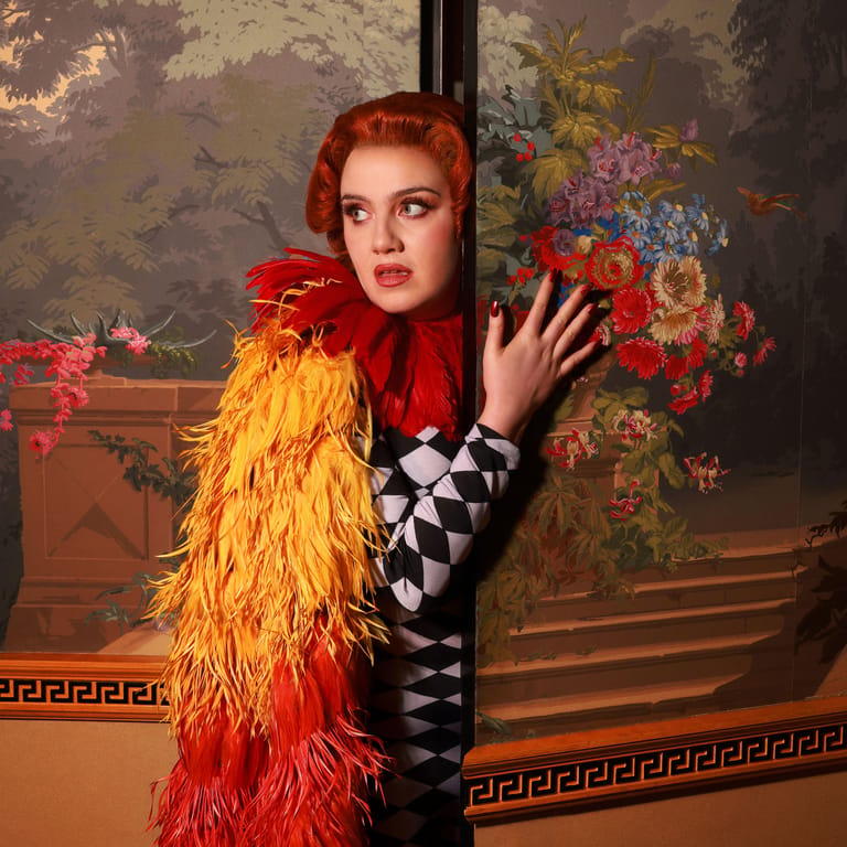 Woman with styled red hair wearing a red headwrap and yellow-to-red feathered boa poses against an ornate floral backdrop.