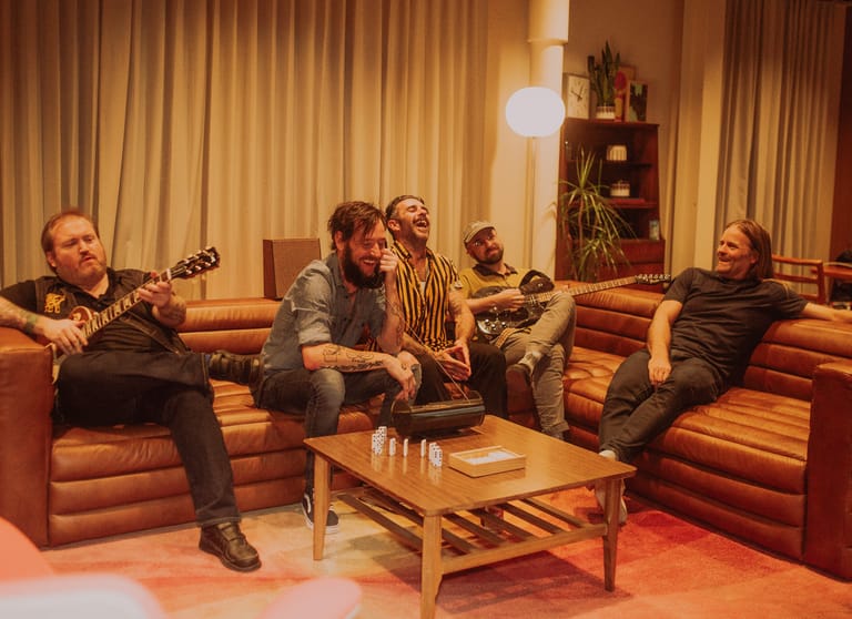 Band members seated on orange couches in a warm-lit retro lounge setting during a casual group discussion.