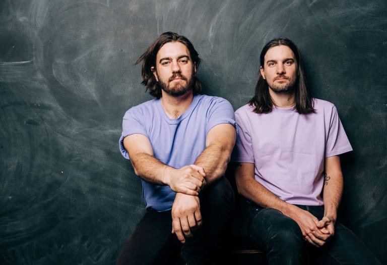 Two men with long dark hair sit side by side against a textured grey backdrop.