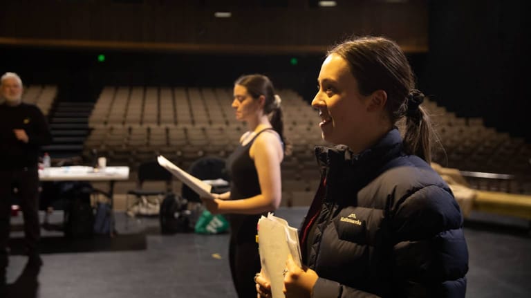 Person in black jacket holding a clipboard on a theatre stage with seated pianist and empty seating visible behind.