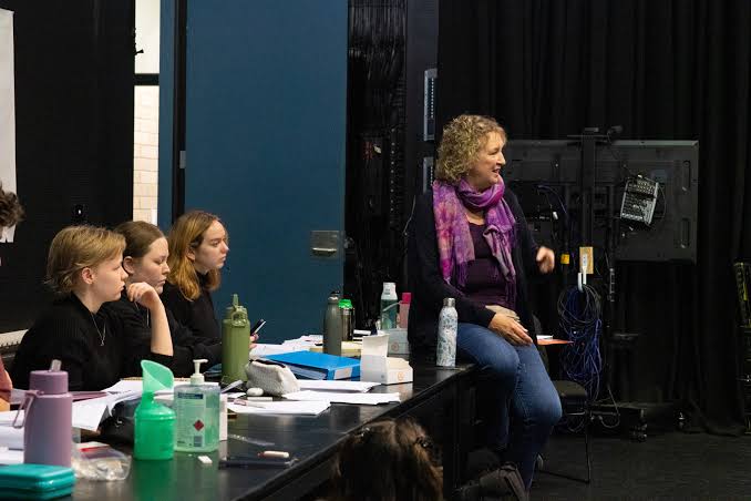 Woman in purple scarf speaks to seated group during workshop or rehearsal in theatre space.