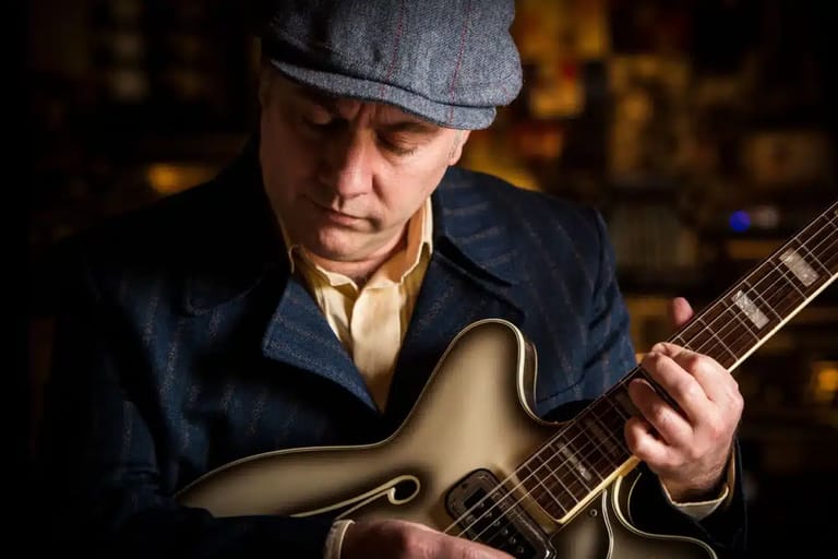 Musician in a grey cap and navy jacket playing an acoustic guitar against a blurred warm-toned background.