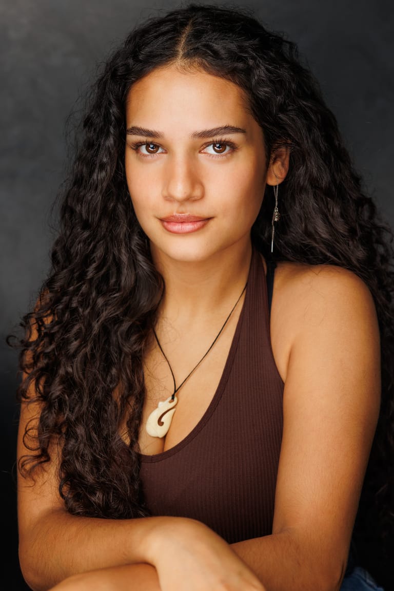Person with long dark wavy hair and shell pendant necklace against neutral grey background.