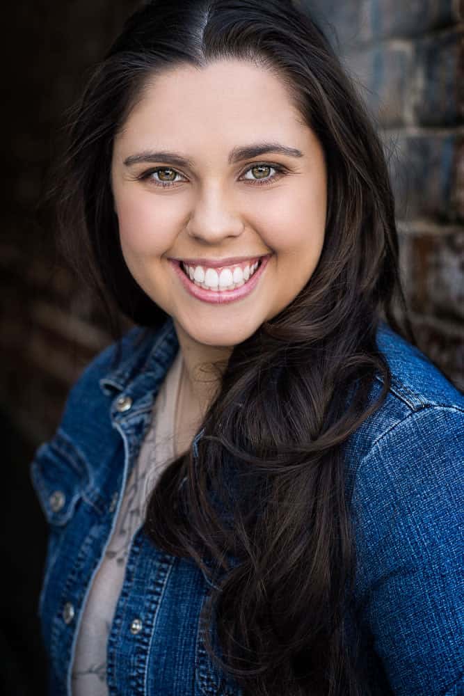 Woman with long dark hair wearing a blue denim jacket, smiling at the camera against a brick wall backdrop.