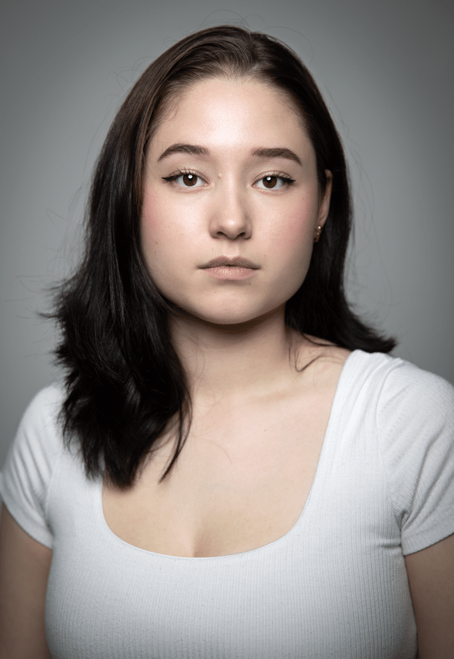 Person with long dark hair wearing a white ribbed top, against a neutral grey background.