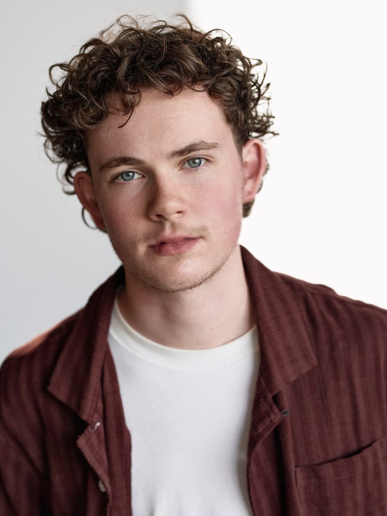Young man with curly dark hair wearing a burgundy shirt over a white t-shirt, against a neutral grey background.