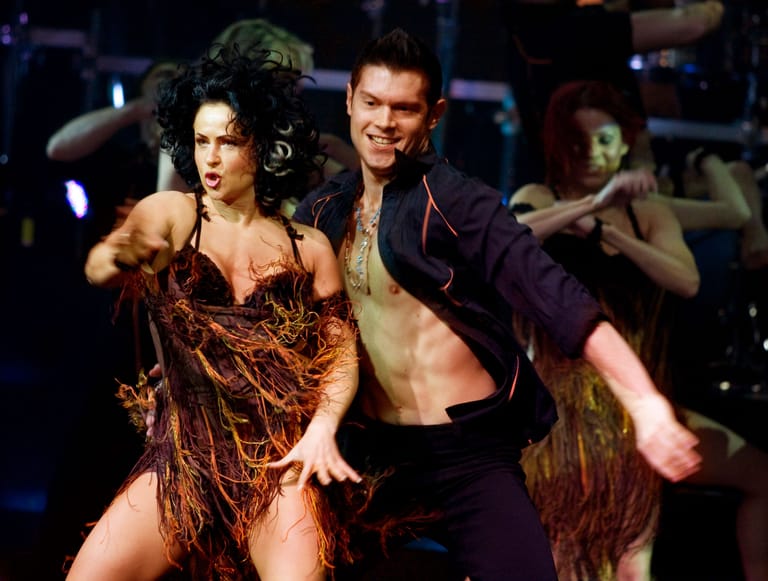 Two performers in fringed costumes dance together on a stage lit by blue spotlights during a musical theatre production.