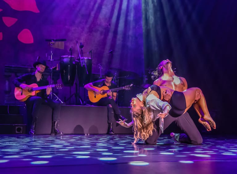 Performer in gold outfit reclines on stage floor whilst musicians with acoustic guitars play beneath purple spotlights.