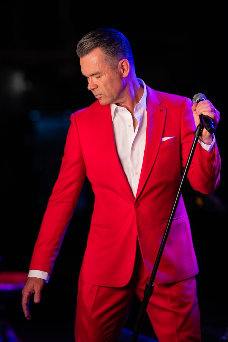 Performer in a bright red suit holding a microphone on stage, lit by red and purple spotlights.