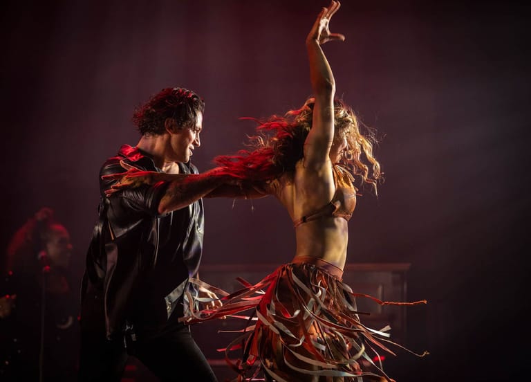 Performer in leather jacket reaches toward shirtless dancer adorned with red fabric and branches against dark stage backdrop.
