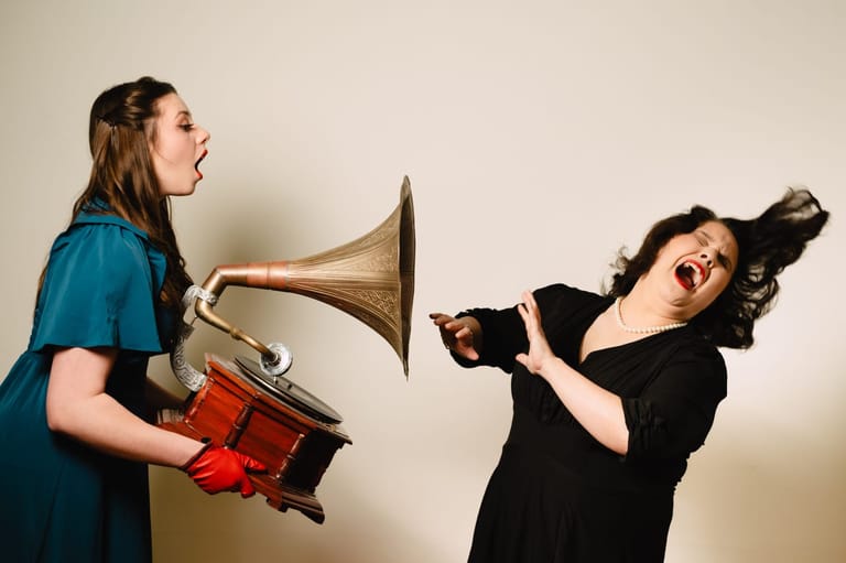 Two performers playfully interact with brass and percussion instruments against a neutral backdrop.