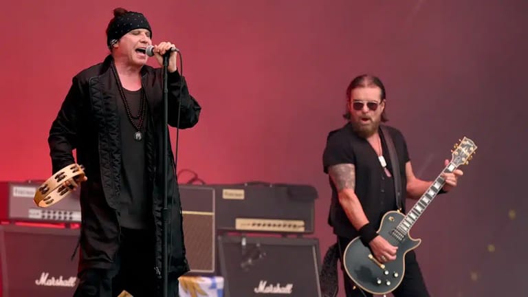 Singer in black suit performing at microphone with guitarist on stage against red lighting.