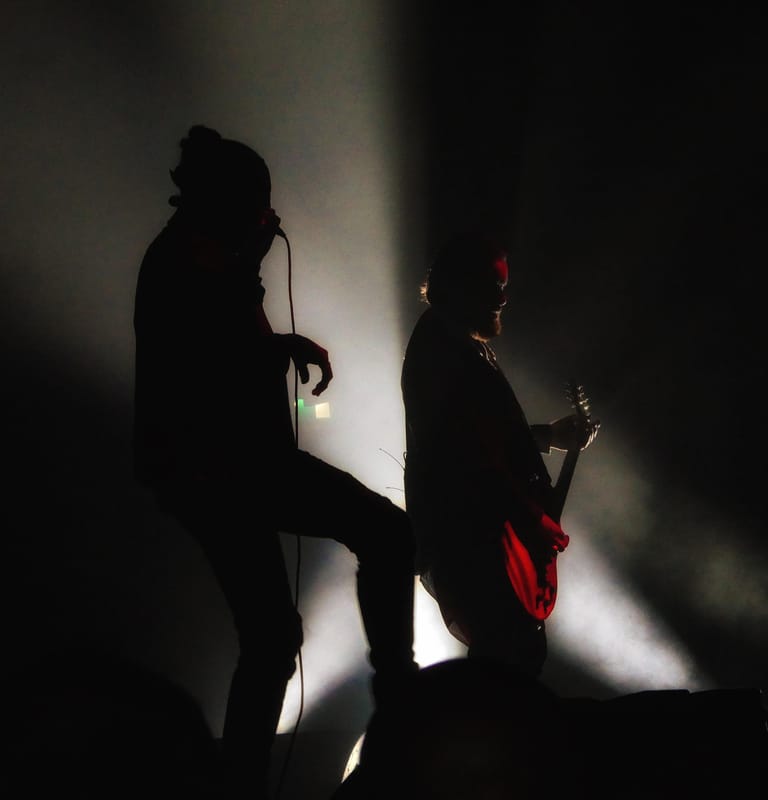 Silhouetted singer and guitarist performing on stage, lit by dramatic white and red stage lighting.