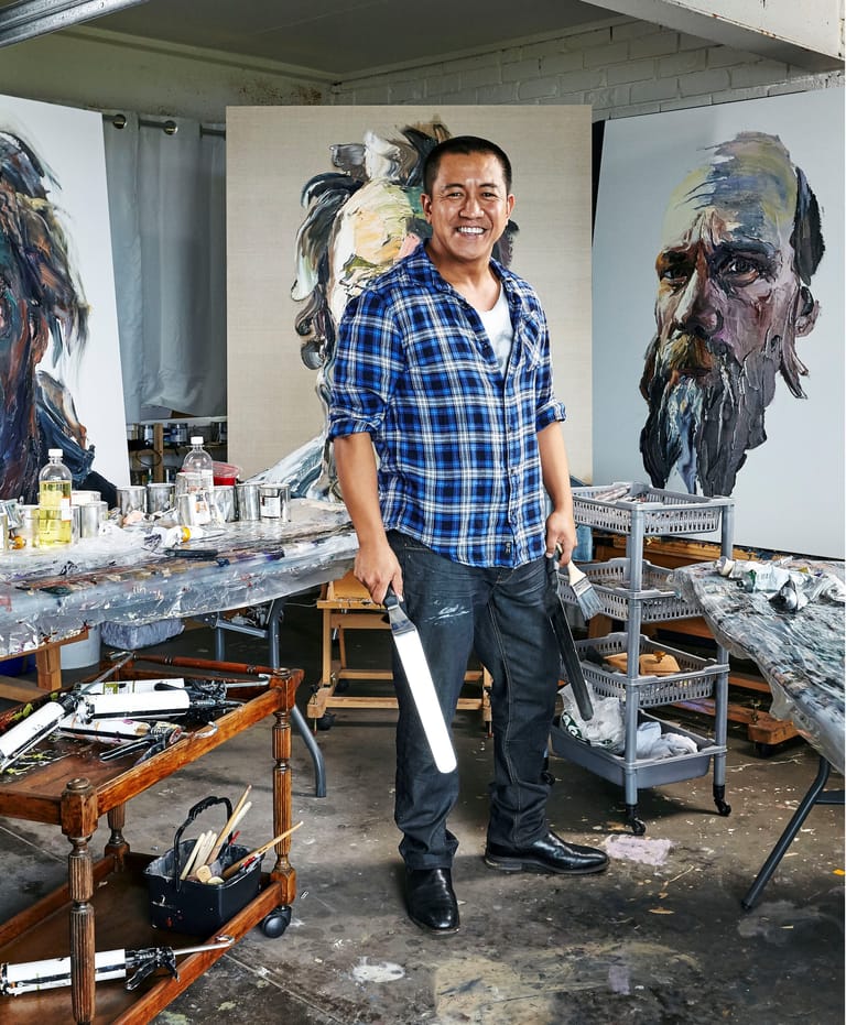 Artist in blue plaid shirt stands in studio surrounded by portrait paintings and art supplies on work table.