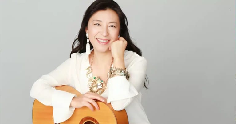 Musician holding an acoustic guitar, wearing a white blazer and jewellery, smiling at the camera against a grey background.