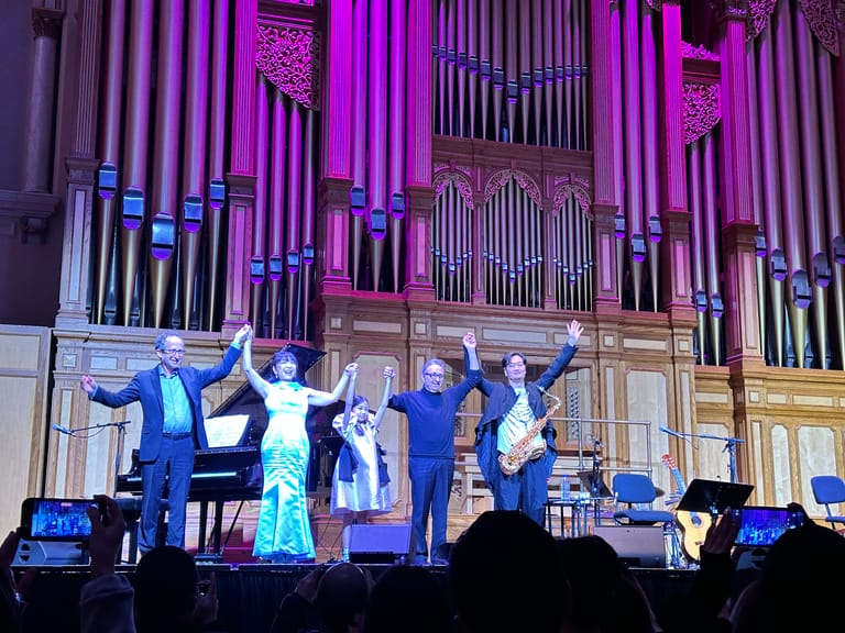 Performer in shimmering dress stands centre stage with band members in blue suits beneath a grand illuminated pipe organ.