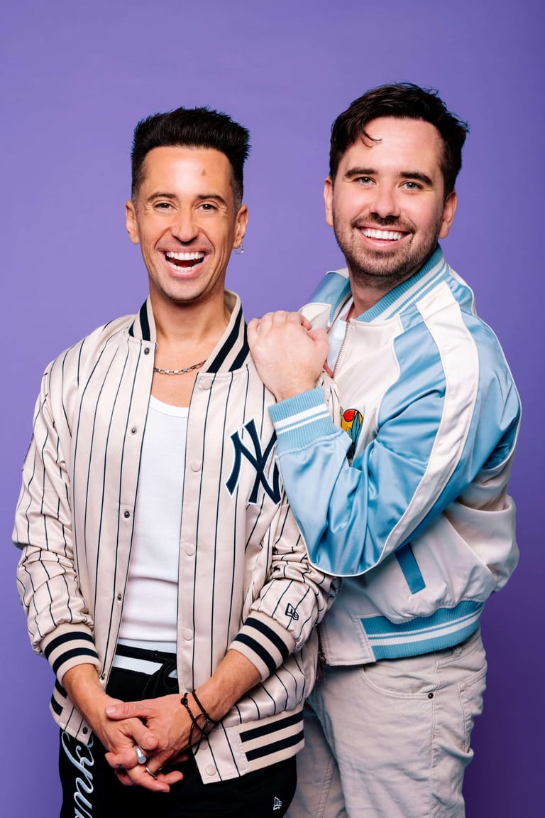 Two smiling men pose together against a purple background, one wearing a pinstriped baseball jacket and the other a blue and white striped jacket.