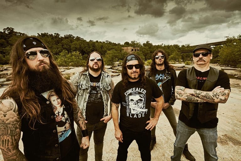 Five band members in dark clothing and sunglasses stand in an arid, sandy landscape under cloudy skies.