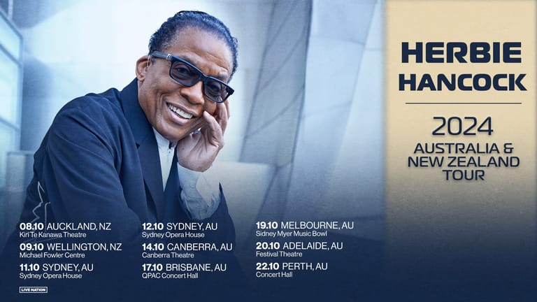 Herbie Hancock wearing sunglasses and a blue suit, smiling while resting his head on his hand against a light blue background.