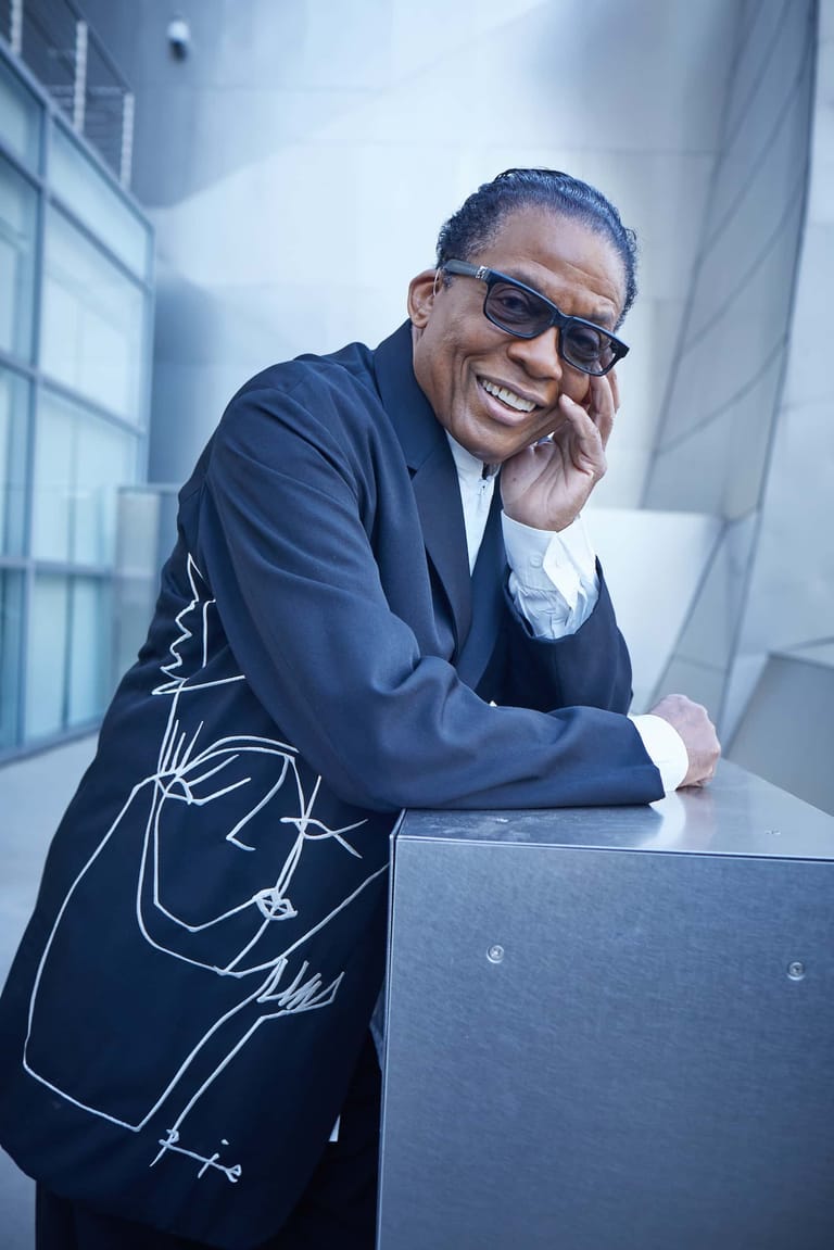 Man in dark suit and sunglasses leans against a table, smiling at camera in a modern office setting.
