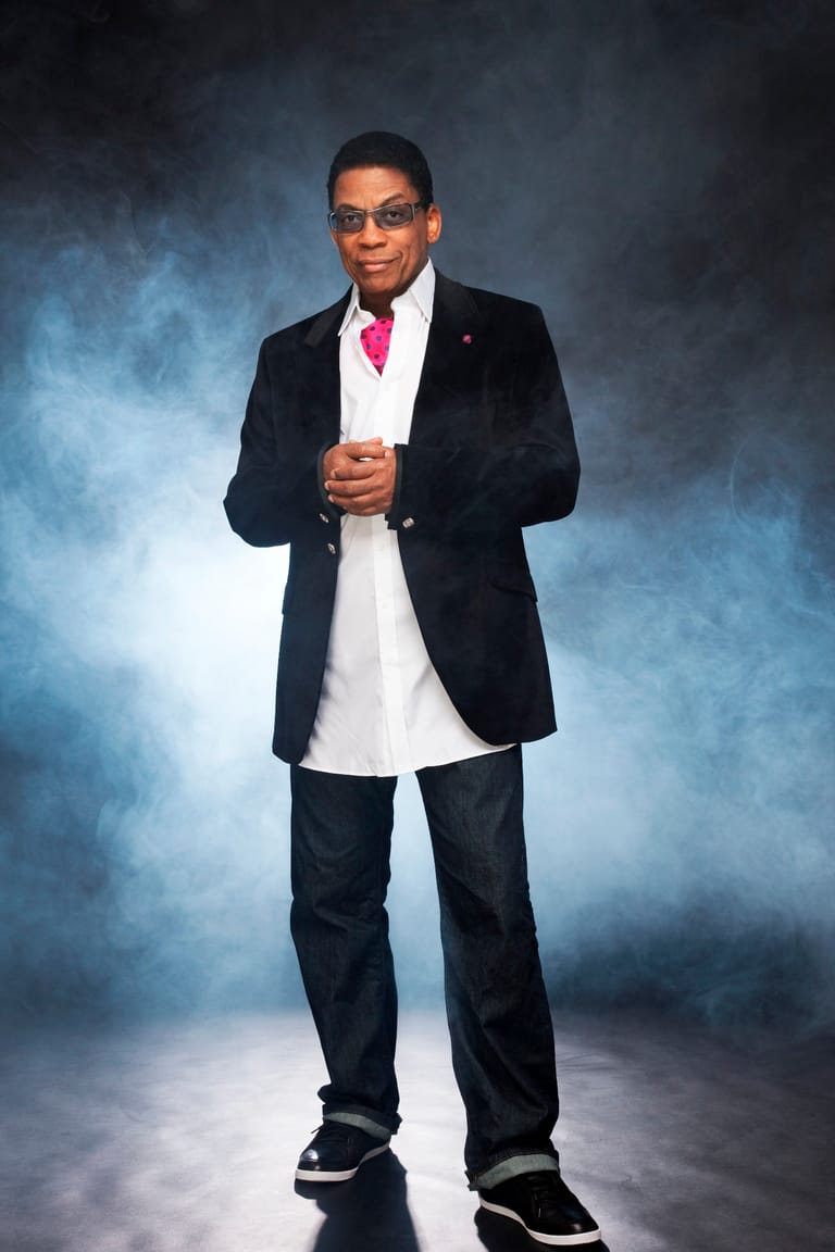 Man in black blazer and pink tie stands against blue-grey studio backdrop, wearing glasses and casual sneakers.