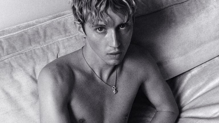 Shirtless person wearing a pendant necklace, posed on a bed in black and white.