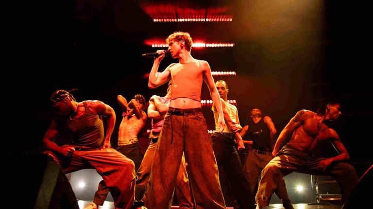 Performer in orange crop top and red trousers singing centre stage, surrounded by band members under warm stage lighting.