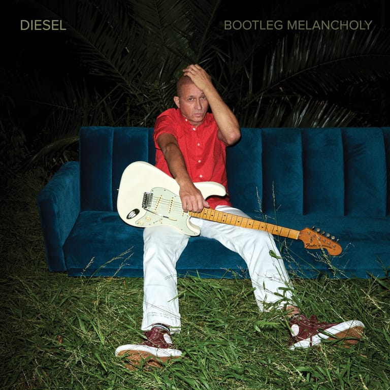 Guitarist in red shirt and white pants holding white electric guitar, seated on blue velvet couch in grass at night.