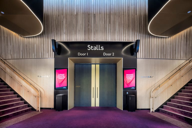 Modern theatre foyer with symmetrical staircase, purple carpeting, and illuminated stalls entrance doors beneath metallic wall panelling.