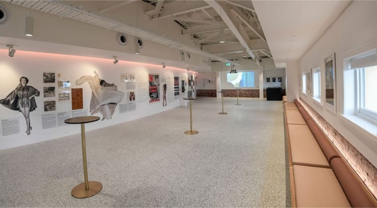 White gallery space with fashion sketches and garments displayed on walls under warm overhead lighting.