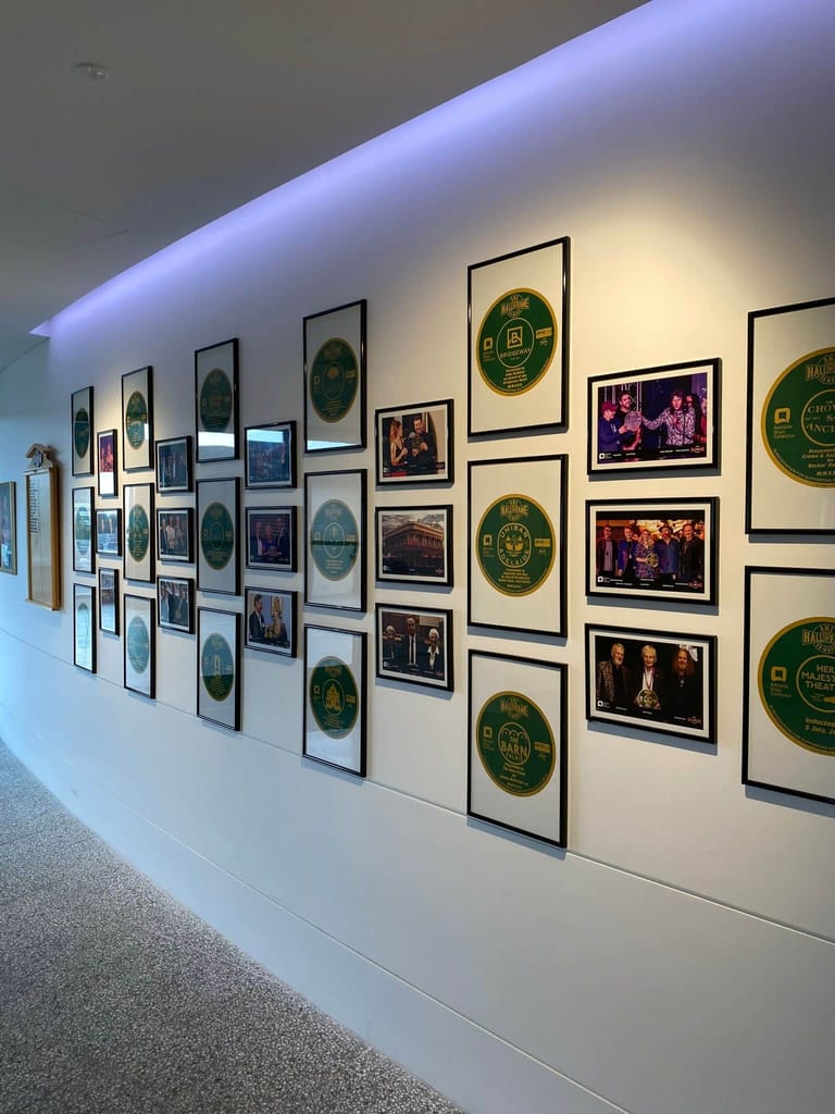 Gallery wall displaying framed photographs and circular green badges under blue and warm overhead lighting.