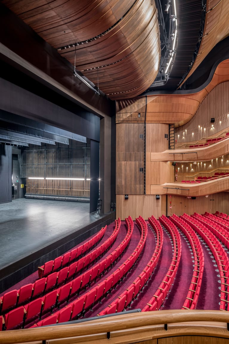 Modern theatre auditorium with curved rows of red seats facing a wooden-panelled stage with contemporary lighting design.