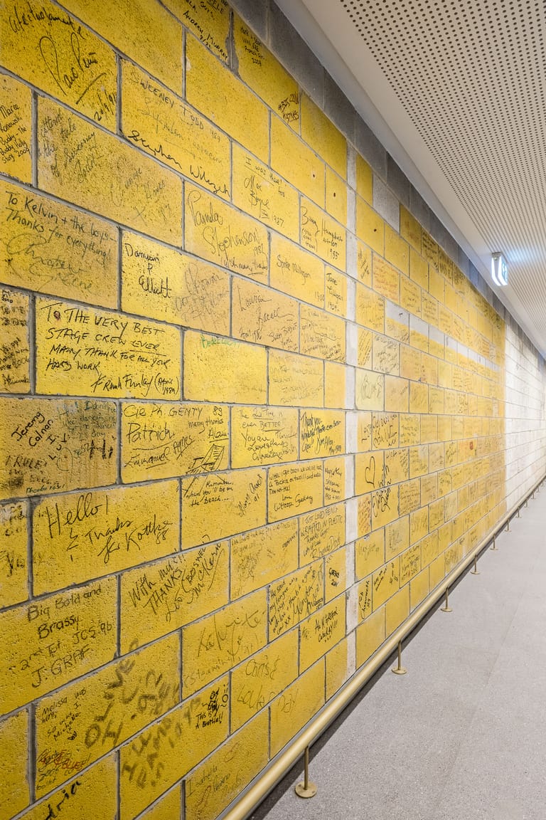 Wall installation of yellow tiles with handwritten messages and signatures covering its entire surface.