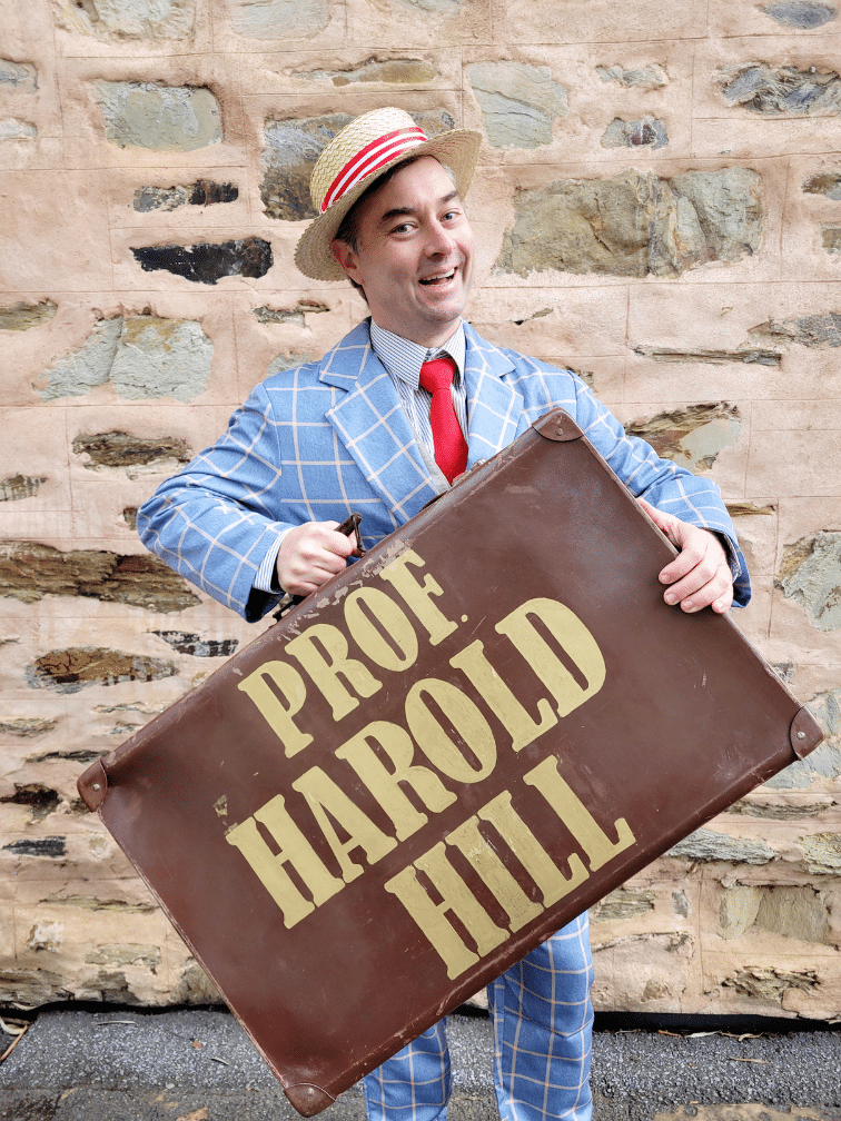 Man in blue checked suit and straw hat holds a vintage "Proof Harold Hill" sign against a stone wall.
