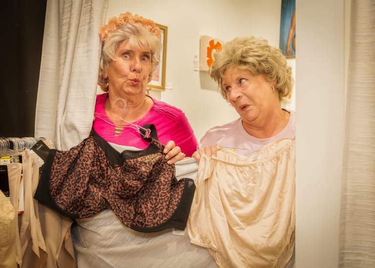 Two performers in colourful costumes examine a patterned garment during a theatre production scene.
